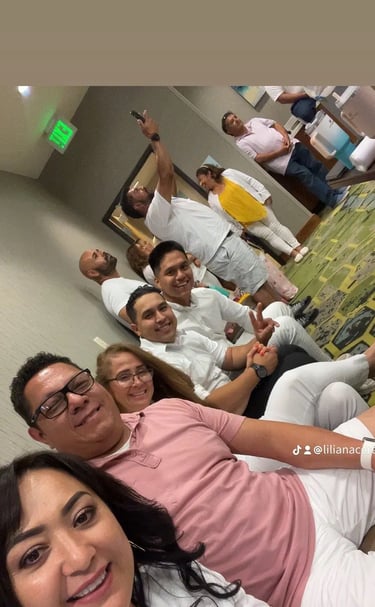 A group of smiling family and friends wearing white and pink shirts gather for a group photo indoors.