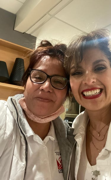 Two smiling women taking a selfie indoors wearing white jackets and casual accessories.
