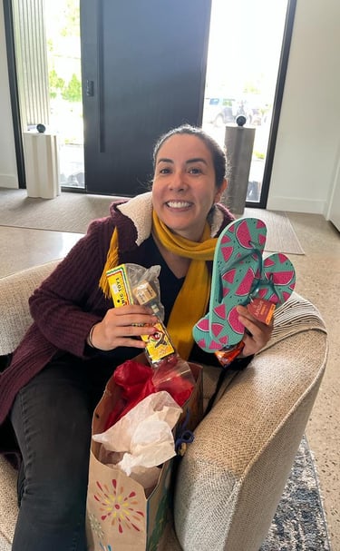 Smiling woman sitting in a chair holding a gift bag, watermelon print flip-flops, and small toys.