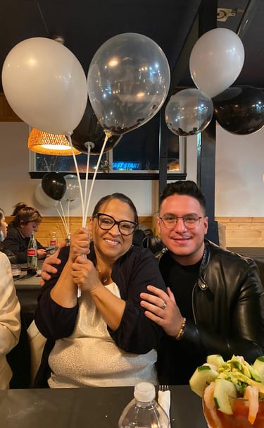 Smiling woman holding white balloons sitting with a man in a leather jacket at a birthday dinner party.