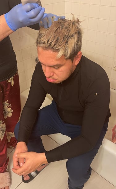 Professional applying blonde hair bleach to a man's short hair in a home bathroom setting.