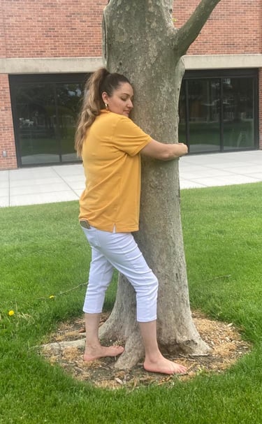 A barefoot woman in a yellow shirt and white pants hugging a large tree on a green lawn.