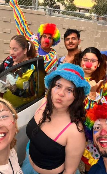 A group of happy friends in colorful clown costumes and wigs taking a fun selfie with bright balloons.