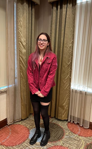 A smiling woman with glasses wearing a red textured jacket, black skirt, and boots standing in a room.