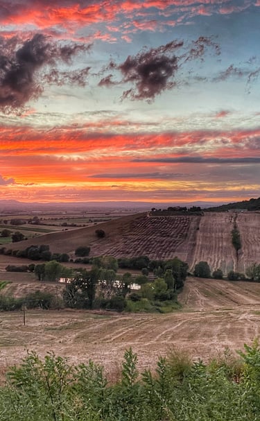 A vibrant sunset over rolling hills and rural farmland in the Italian countryside.