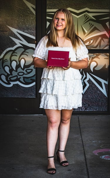 A smiling blonde graduate in a white tiered dress holds a red diploma, celebrating her achievement!