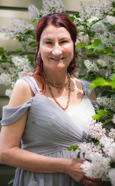 Smiling woman with auburn hair in a grey dress posing by white lilac bushes.