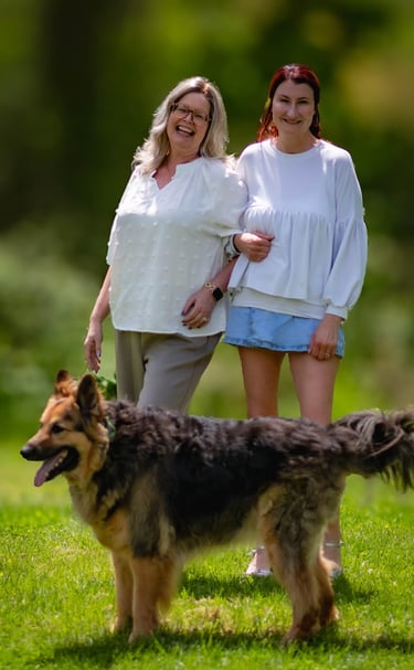 Two smiling women standing together in a sunny park with their large long-haired German Shepherd dog.