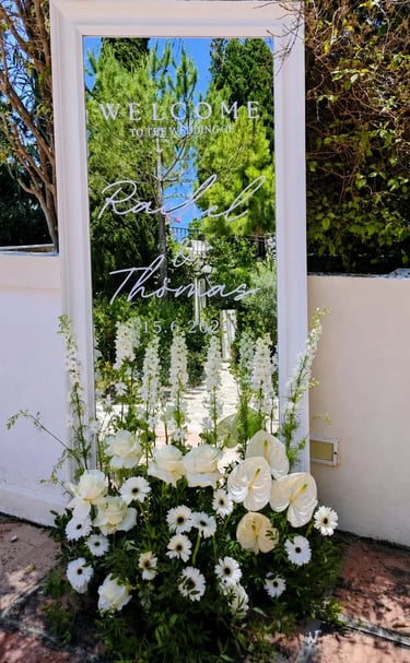 A white-framed mirror wedding welcome sign for Rachel and Thomas decorated with white flowers.
