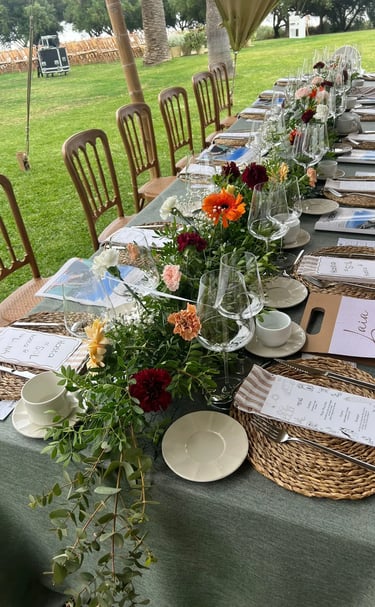 Long outdoor banquet table set with green linen, woven placemats, and a colorful floral centerpiece.