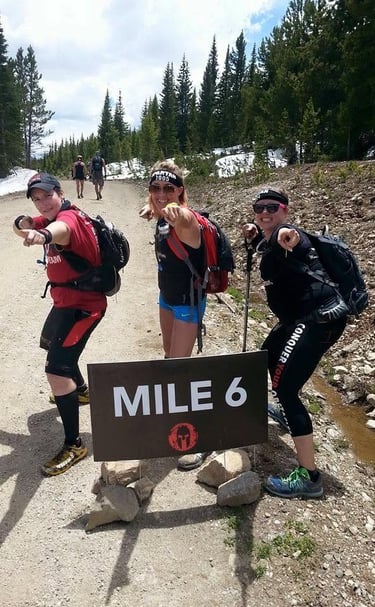 Three Spartan Race athletes posing by a Mile 6 sign on a mountain hiking trail with pine trees.