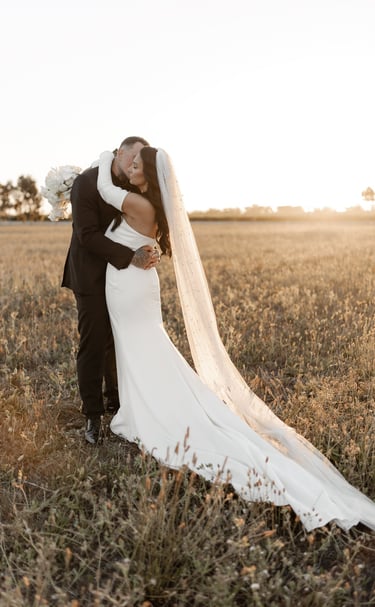 a bride and groom kissing in a field at sunset