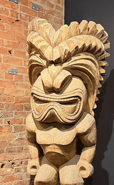 Hand-carved wooden Tiki statue with intricate facial details against a red brick wall.
