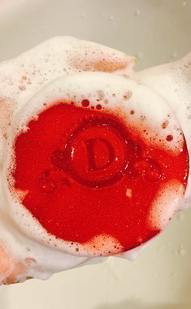 A round red bar of soap with a letter D engraving held in a hand covered in white soapy bubbles.