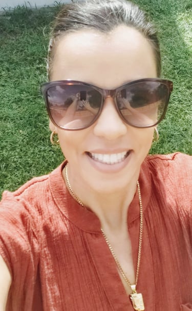 Smiling woman wearing tortoise shell sunglasses and gold jewelry in a green garden.