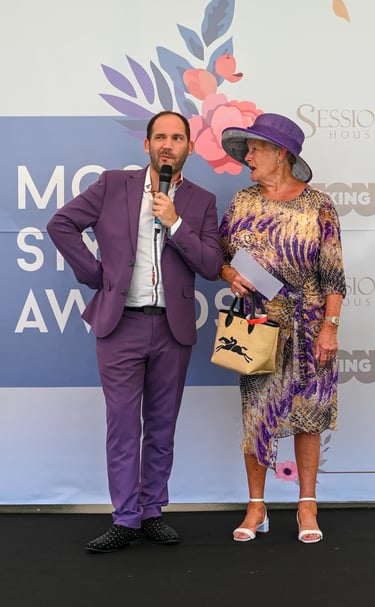 Alex in purple suit with well-dressed lady in purple hat
