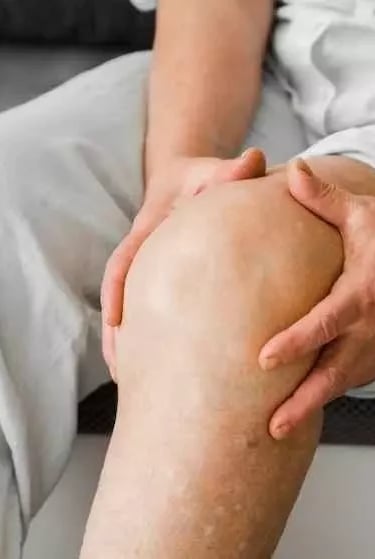 Homem mais velho sentado na cama com ambas as mãos na região do joelho