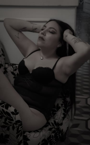 Una mujer con tatuajes en los brazos, vestida con lencería negra de encaje, posando en un sillón