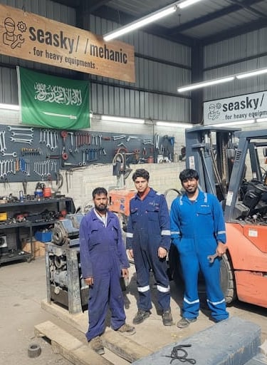 Heavy equipment mechanics in blue coveralls pose in a workshop near a forklift and tool wall.