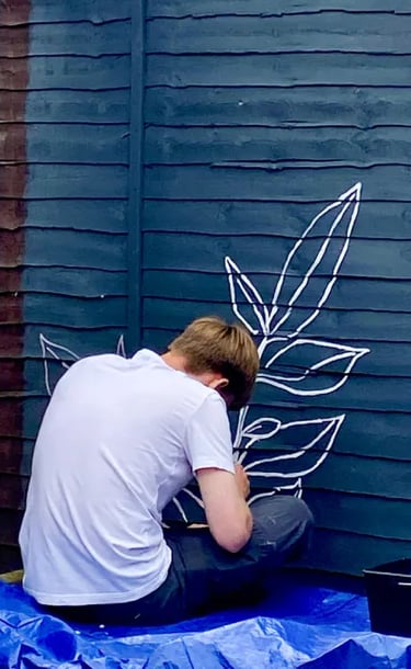 Muralist Byron sits on floor, painting graphic plant on grey wooden garden fence.