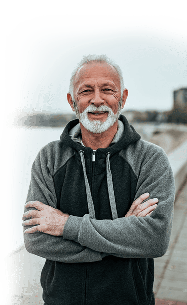 A smiling senior man with a white beard wearing a grey hoodie while standing outdoors.
