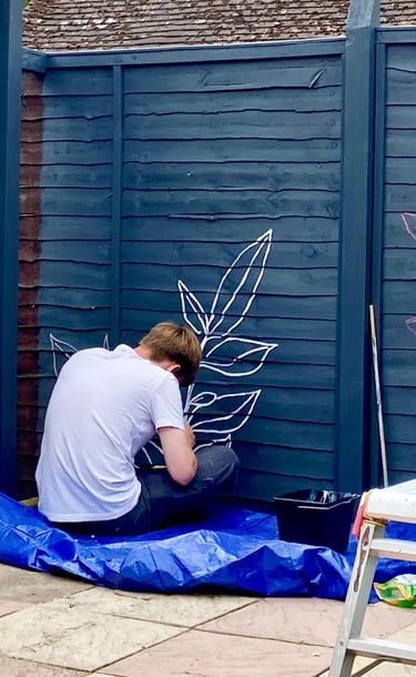 Muralist Byron sits on floor, painting graphic plant on grey wooden garden fence.