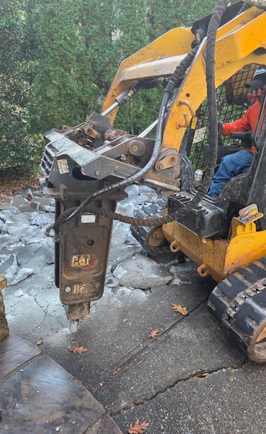 Yellow Cat skid steer with hydraulic hammer attachment breaking up a concrete driveway for demolition.