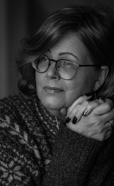 Black and white portrait of a thoughtful, mature woman with eyeglasses, resting her chin on clasped hands.