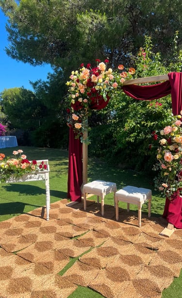 Outdoor wedding ceremony setup with a floral wooden arch, red drapery, two white stools, and a woven rug on the grass.