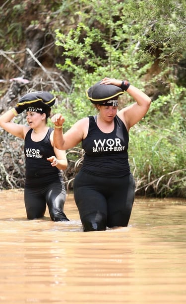 Two women participating in a Spartan Race obstacle course carry sandbags while wading through muddy water.