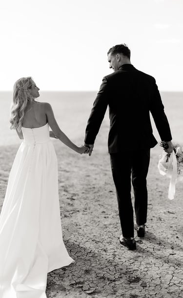 a bride and groom holding hands and walking down a beach