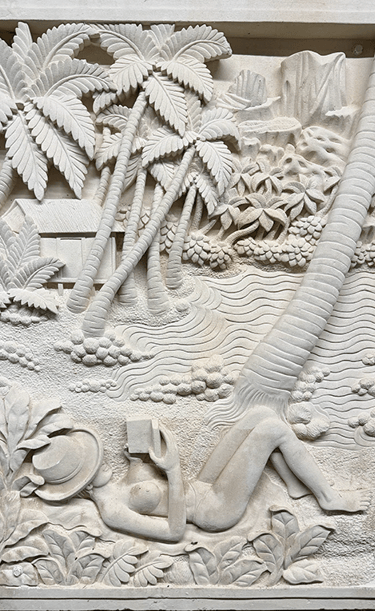 Hand-carved stone relief showing a woman reading under palm trees on a tropical beach.