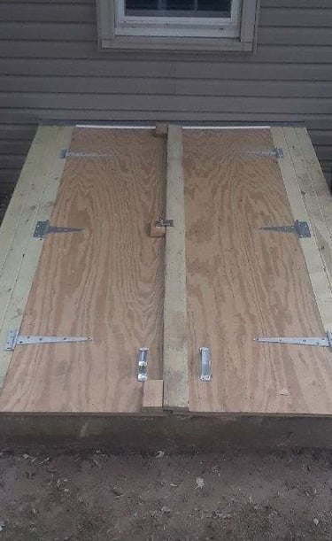 Custom-built wooden storm cellar doors made of plywood and lumber with metal hinges outside a home.