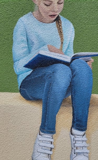 Fresque peinte sur façade. Une fille lit assise sur le bord d'un muret.