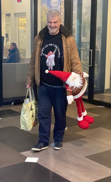 Willem in kersttrui met een kerstman en boodschappentas, lachend in de gang van een gebouw