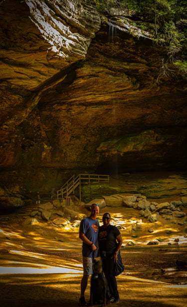 A couple with a dog standing inside Ash Cave at Hocking Hills State Park under a massive rock overhang.