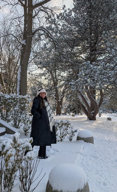 A woman in a long black winter coat and scarf stands in a snow-covered park surrounded by frosted pine trees.