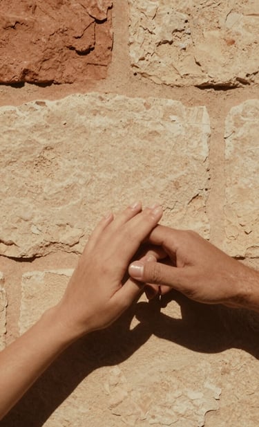 A cinematic, vertical frame capturing a couple's hands gently touching against a background of a sun-bleached stone wall. The style is minimalist and emotive, focusing on textures. The warm sandy tones of #FDF8F0 dominate the scene, with deep terracotta #AD7B5B shadows providing depth and an authentic, timeless feel.
