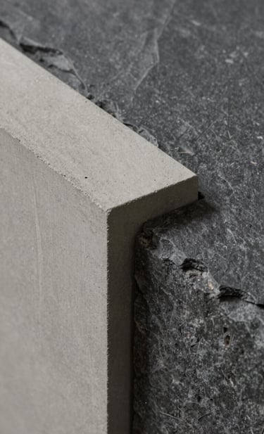 A detailed close-up photograph of architectural materials. A slab of light neutral grey concrete meets a textured dark slate grey stone. The lighting is soft and directional, focusing on the tectonic precision and the low-impact materials used by Estúdio Roda.