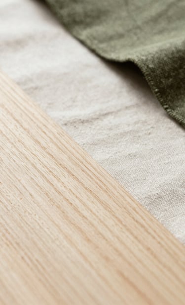 A macro photograph of natural textures used in sustainable design, such as light-toned wood grain and organic linen fabric. The composition is artistic and minimalist, shot in a bright, airy environment in South America. The palette focuses on off-white and muted olive green.