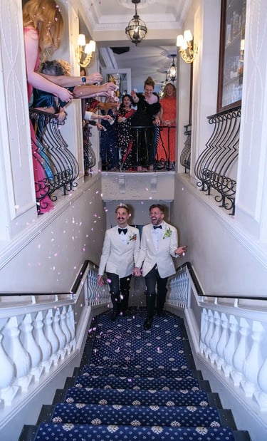 Grooms in white tuxedo jackets walking down stairs as guests throw pink wedding confetti.