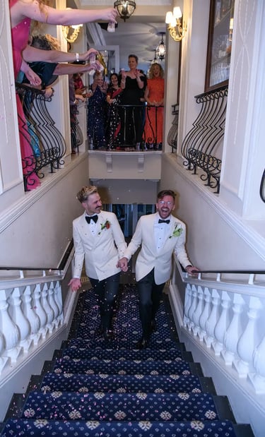 Grooms in white tuxedo jackets celebrate their wedding walking down a grand staircase while guests throw confetti.