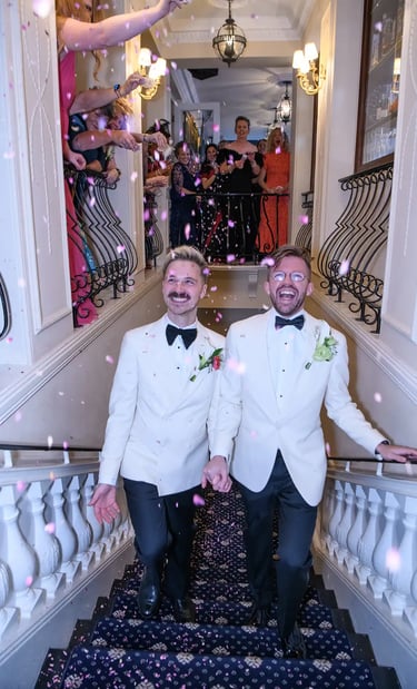 Two grooms in white tuxedo jackets walking down a staircase as guests throw pink flower petal confetti.