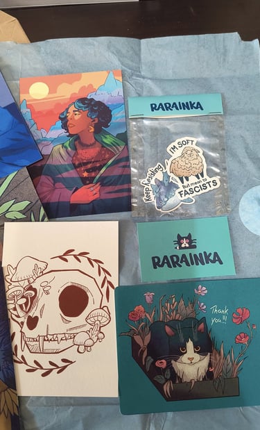 A collection of Rarainka art prints and stickers featuring a black cat, floral skull, and diverse character illustrations.