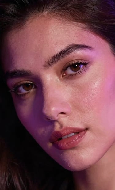 Close-up photograph of an adult model looking confidently into the camera, glowing under soft purple and pink neon highlights, deep shadows, luxurious global aesthetic, Latin American / Global style.