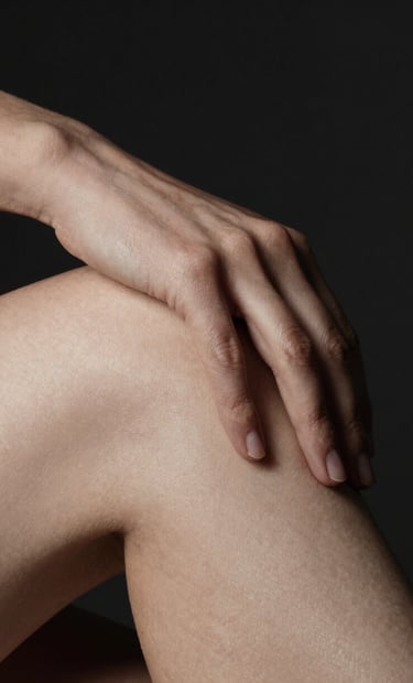 A minimalist macro photograph focusing on the soft texture of skin where a hand rests upon a knee. The lighting is extremely low-key, with a single soft light source from the side creating a gentle highlight in off-white against deep charcoal and dark black shadows. North American / US studio setting.