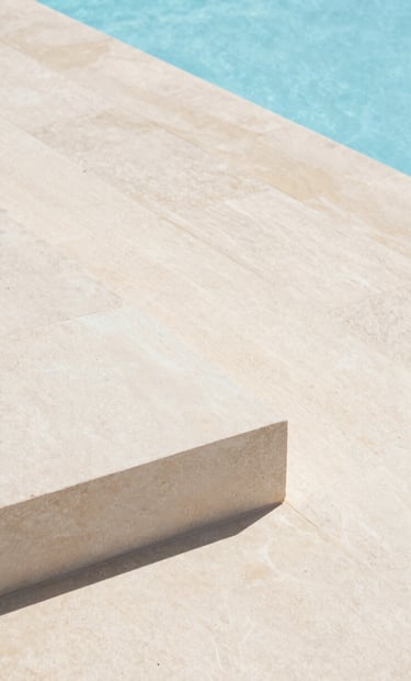 A close-up architectural detail of a minimalist stone terrace in Portugal. Off-white stone slabs meet the edge of a light blue infinity pool. Precise lines, clean composition, bright daylight.