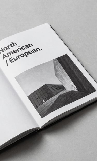 A high-angle shot of a minimalist art book lying open on a gray surface, displaying black and white architectural photography, clean typography visible on the page, North American / European.