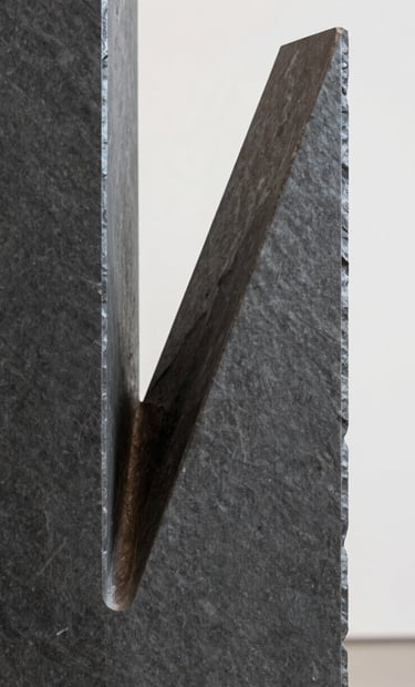 Close-up detail of a contemporary sculpture made of dark slate gray polished metal, reflecting soft light. The composition is minimalist and focuses on the interplay of sharp edges and curves. Professional studio lighting in a Latinoamericano / Español gallery setting.