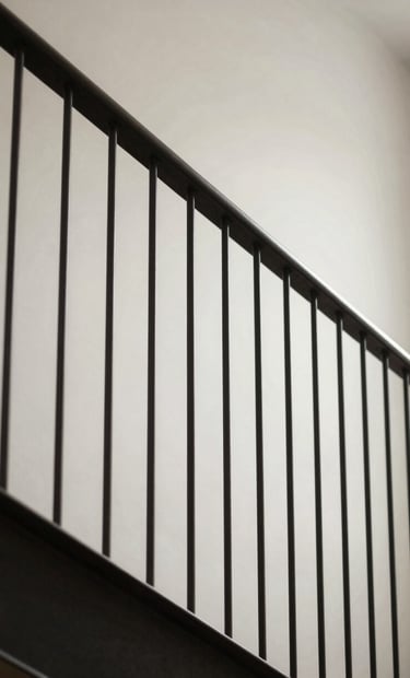 A low-angle shot of a minimalist staircase railing made of deep charcoal steel. The light hits the slim vertical spindles, creating sharp shadows against a soft off-white wall in a modern Nordic interior.